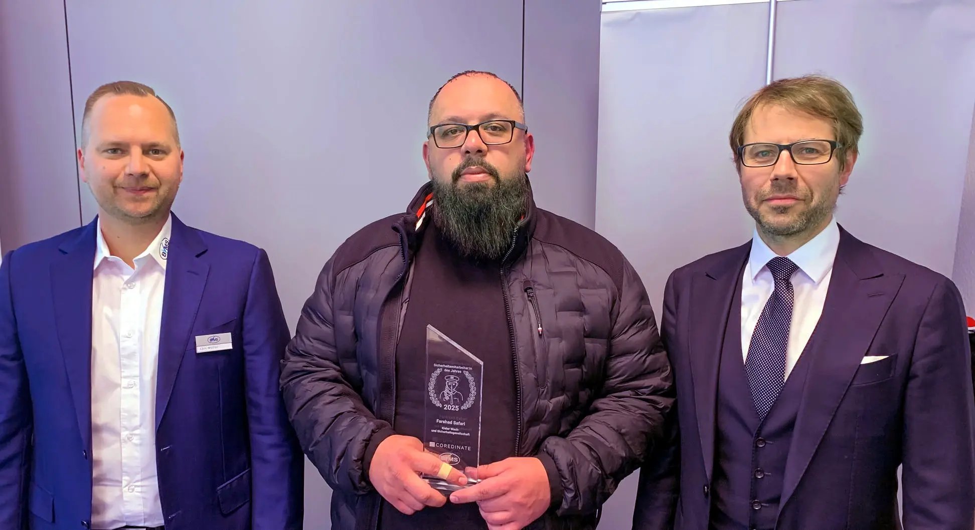 Farshad Safari se tient entre Lars Müller (à gauche) et Michael Kulig (à droite) avec son prix d'« agent de sécurité de l'année 2025 » et tient le trophée dans ses mains.