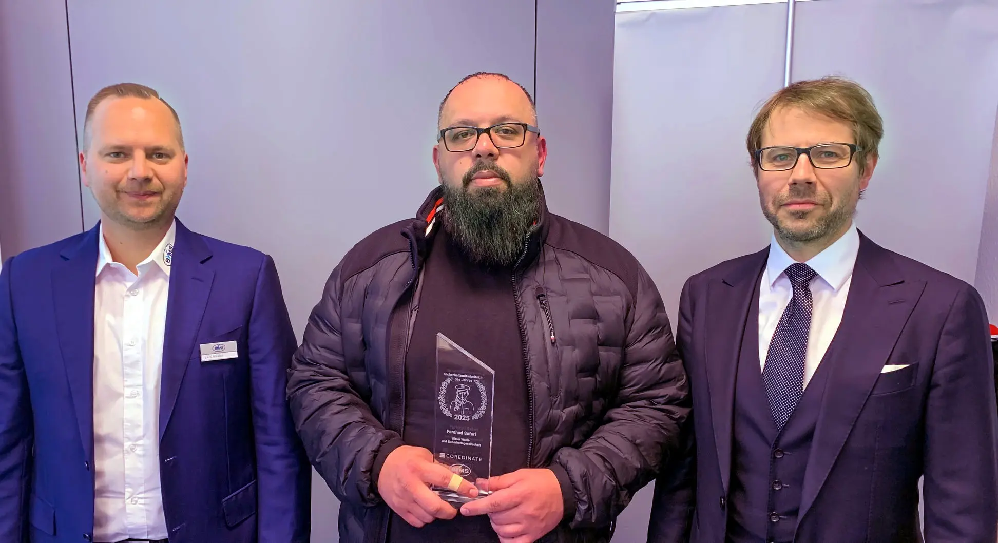 Farshad Safari steht mit seiner Auszeichnung als „Sicherheitsmitarbeiter des Jahres 2025“ zwischen Lars Müller (links) und Michael Kulig (rechts) und hält die Trophäe in den Händen.